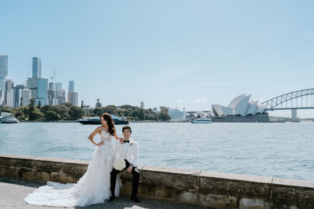 Bride and groom pose by Sydney Harbour with Opera House and Harbour Bridge backdrop on sunny wedding day.