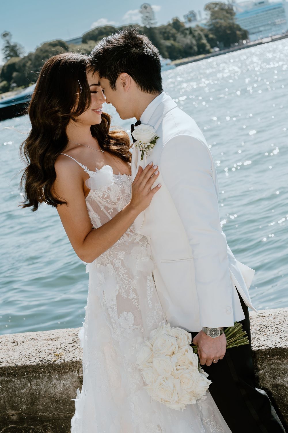 A bride and groom share a romantic moment by a sparkling waterfront, she in lace gown holding white roses, he in white tuxedo.