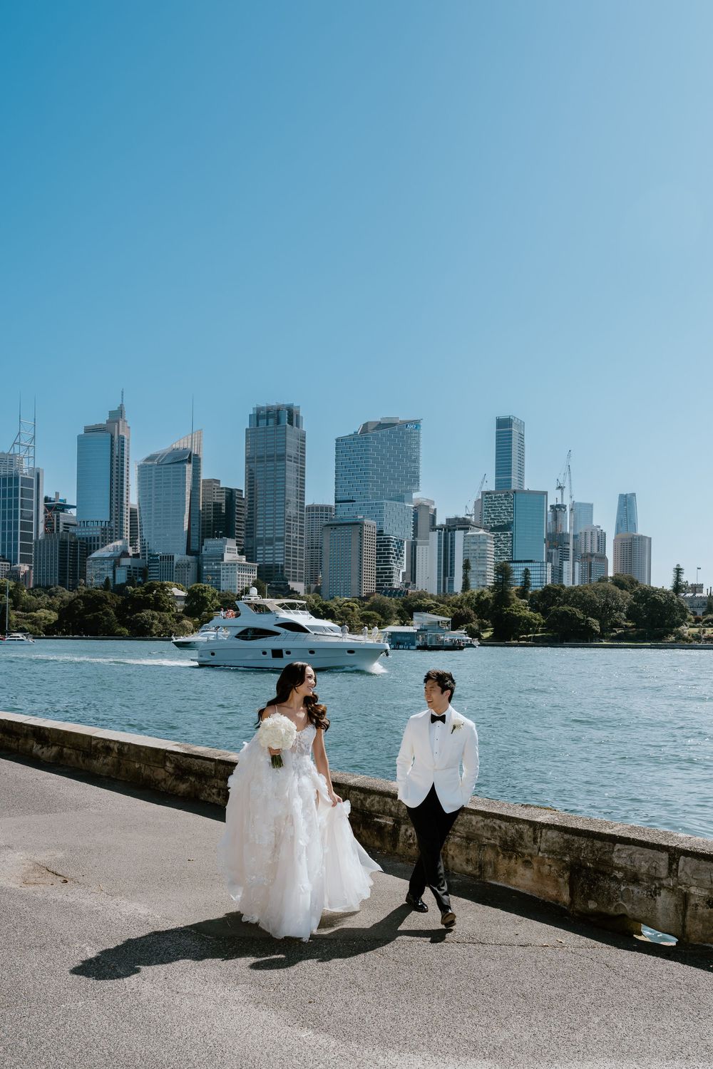 Bride and groom walk along Sydney Harbour waterfront with stunning city skyline and yacht in background on sunny wedding day.
