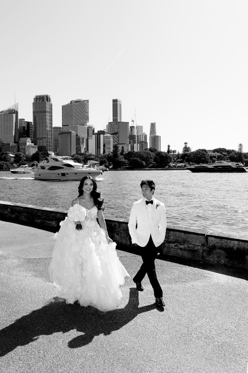 Black and white photo of bride and groom walking by Sydney Harbour waterfront with city skyline backdrop.
