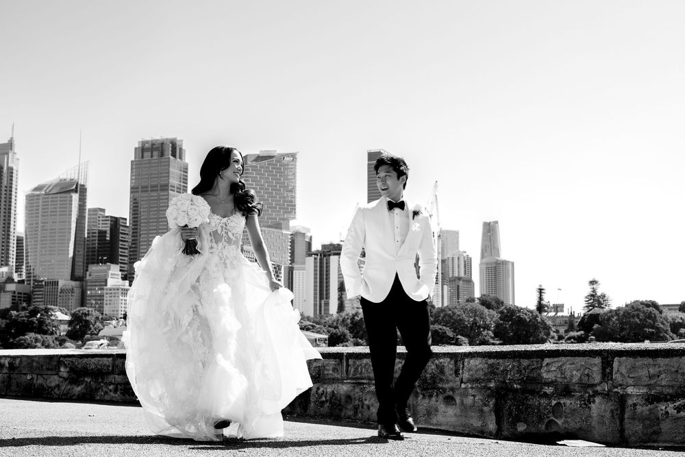 Bride and groom walk together with Sydney city skyline backdrop in elegant black and white wedding portrait.