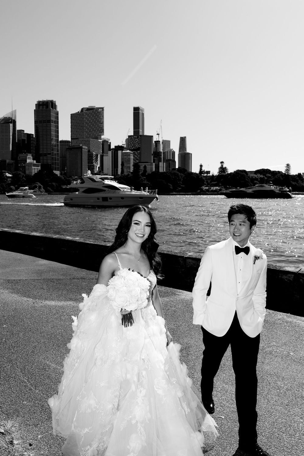 Black and white photo of a bride and groom posing by Sydney Harbour waterfront with city skyline and boats in background.