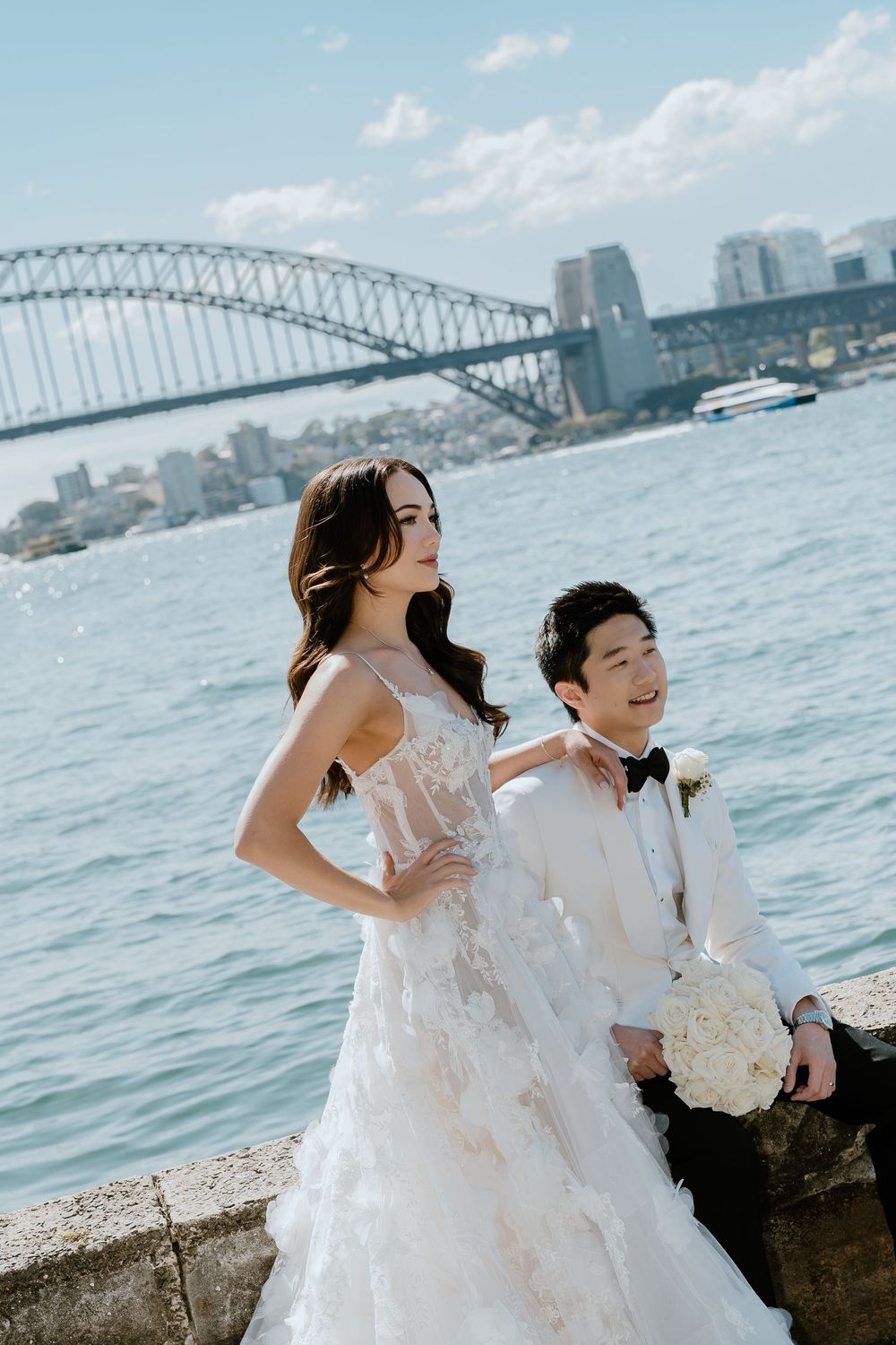 Bride in lace gown and groom in tuxedo pose by Sydney Harbour with iconic Harbour Bridge backdrop on wedding day.