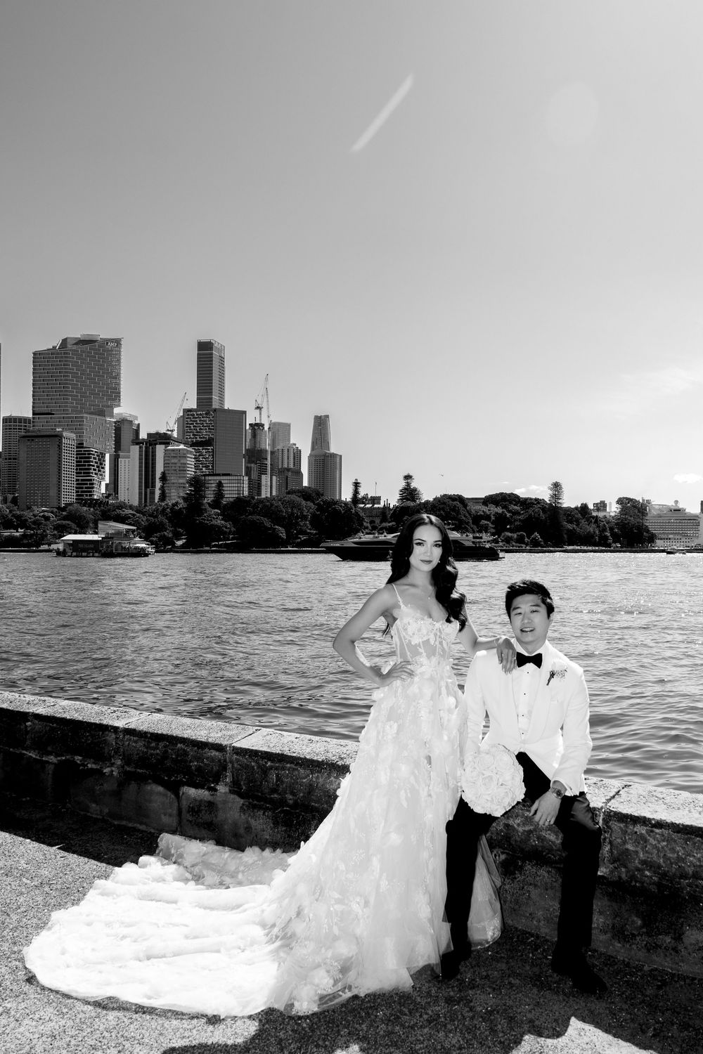 Black and white wedding photo of couple posing by waterfront with city skyline, bride in elegant gown, groom in white tuxedo.