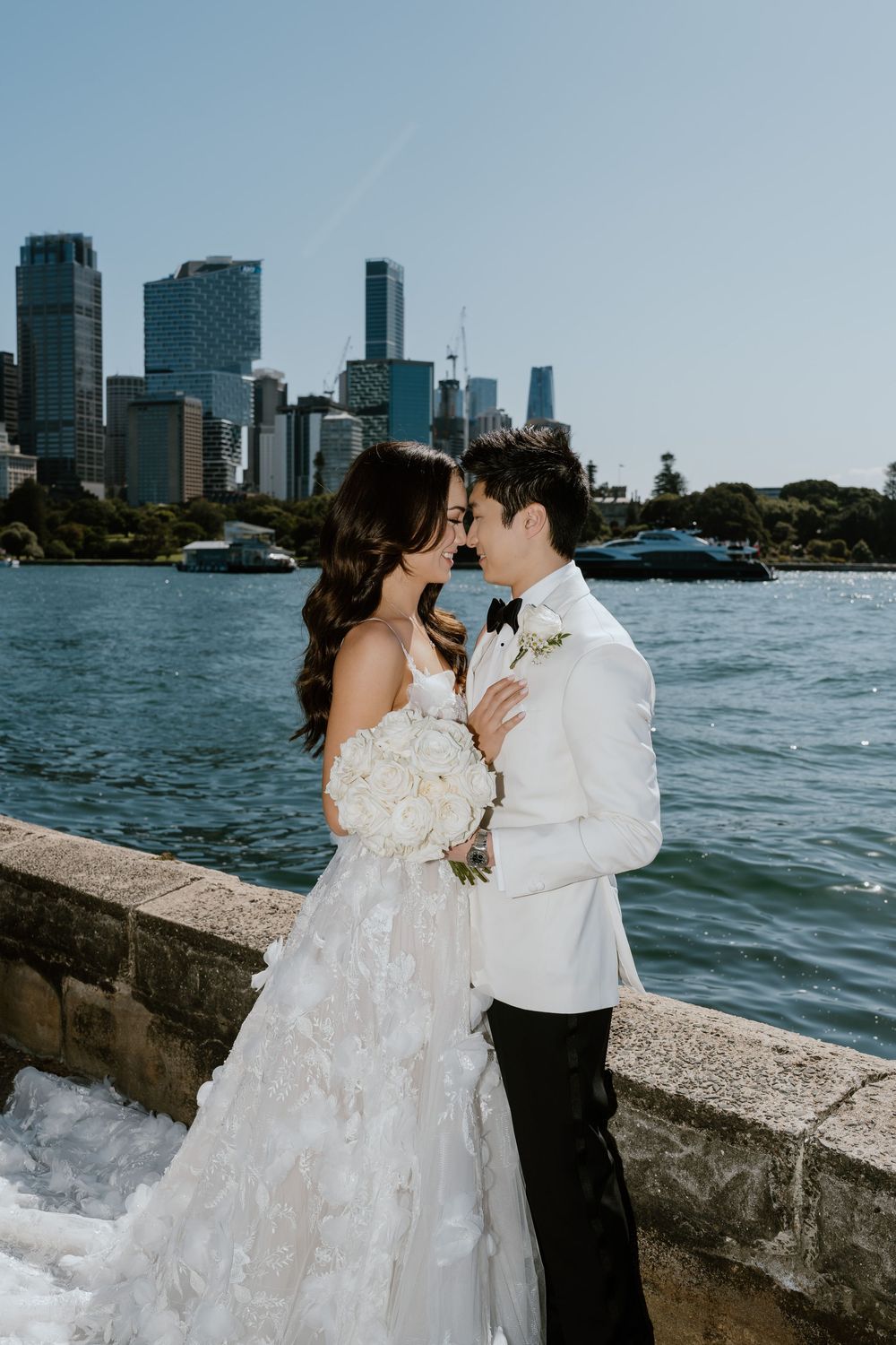Bride and groom share a romantic moment by Sydney Harbour waterfront, with stunning city skyline backdrop on a sunny day.