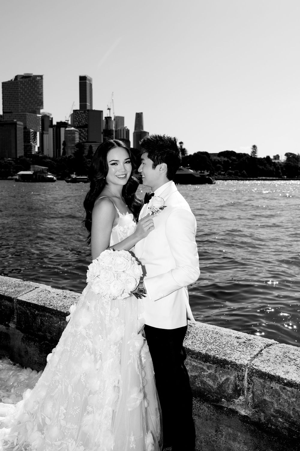 Black and white photo of a bride and groom embracing waterfront with city skyline backdrop on their wedding day.