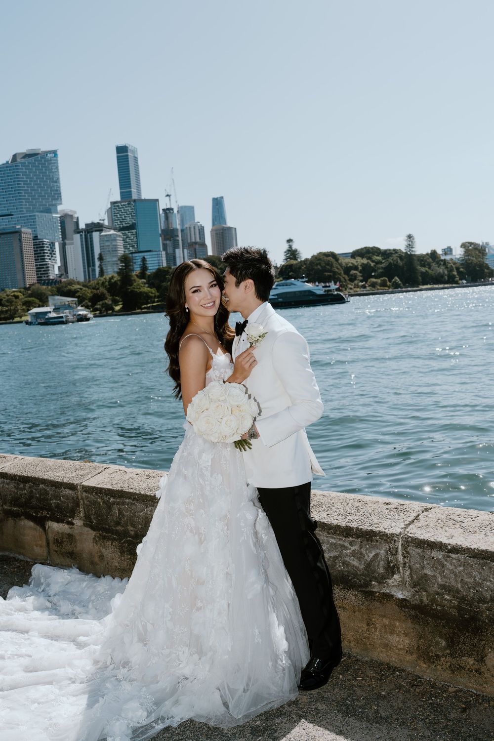 Bride and groom pose by Sydney Harbour waterfront, she holds white bouquet in ruffled gown, he wears white tuxedo jacket.