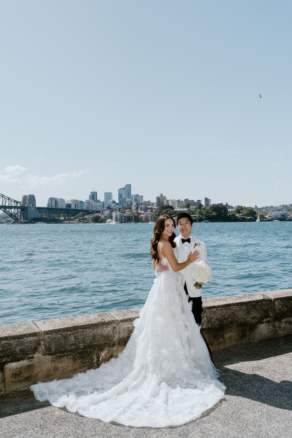 Bride in white gown and groom in tuxedo pose by Sydney Harbour with bridge views on their wedding day.