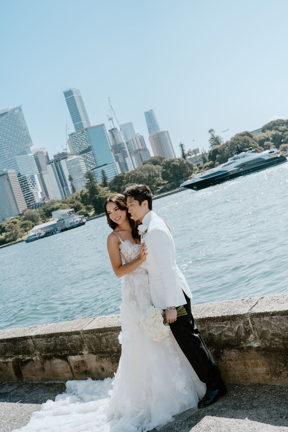Elegant couple embrace waterside with Sydney CBD skyline backdrop during their wedding photo session.