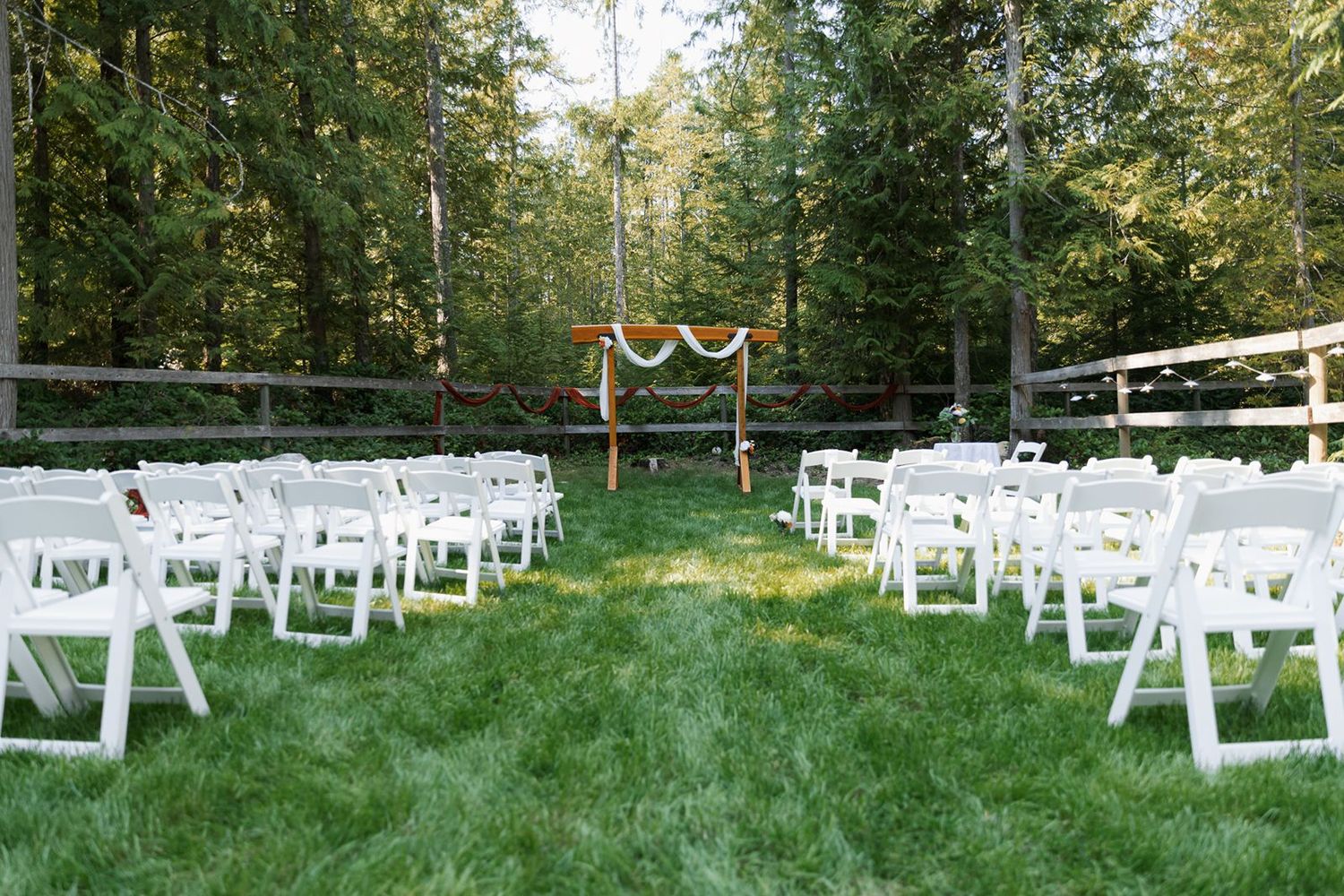 Outdoor wedding ceremony setup with white folding chairs and wooden arch in forest.