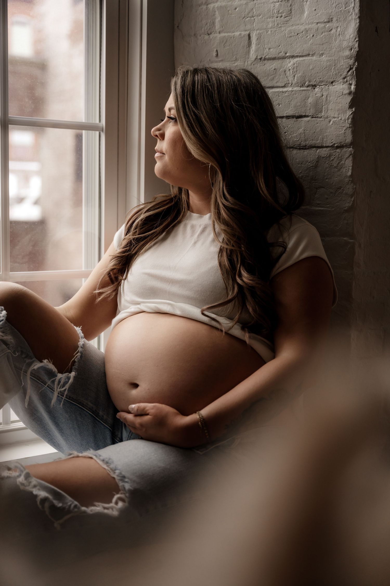 Pregnant woman in white crop top and ripped jeans sitting on windowsill, cradling baby bump while gazing outside.