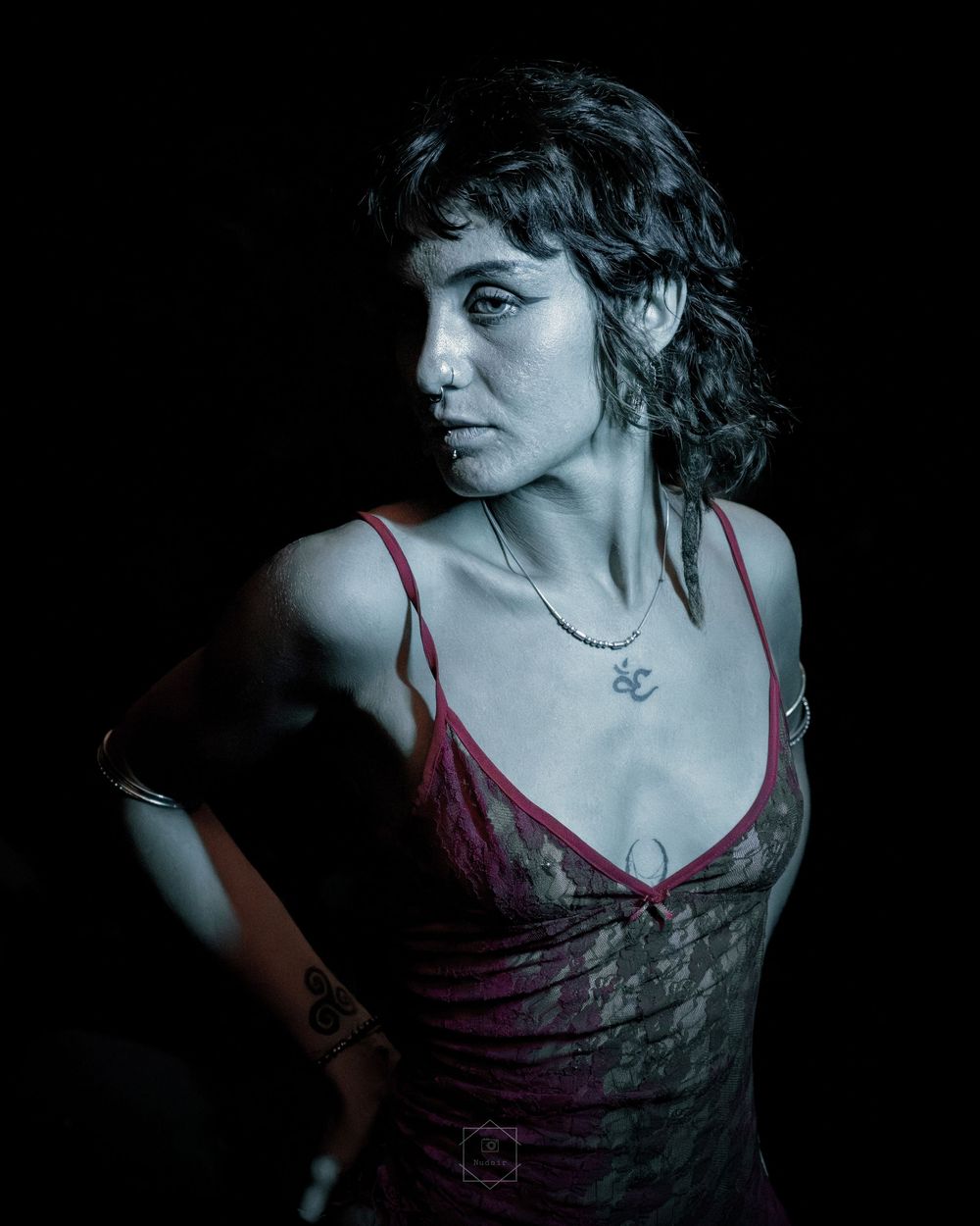 Woman in red lace dress with tattoos and necklace poses against dark background.