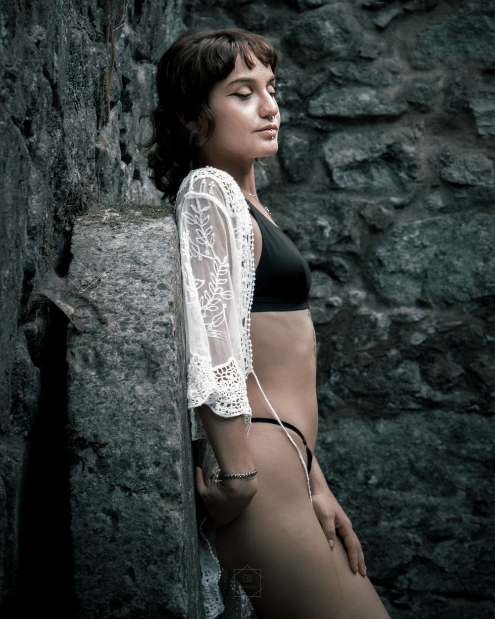 Young woman in black bikini and white lace cover-up posing against a rustic stone wall outdoors.