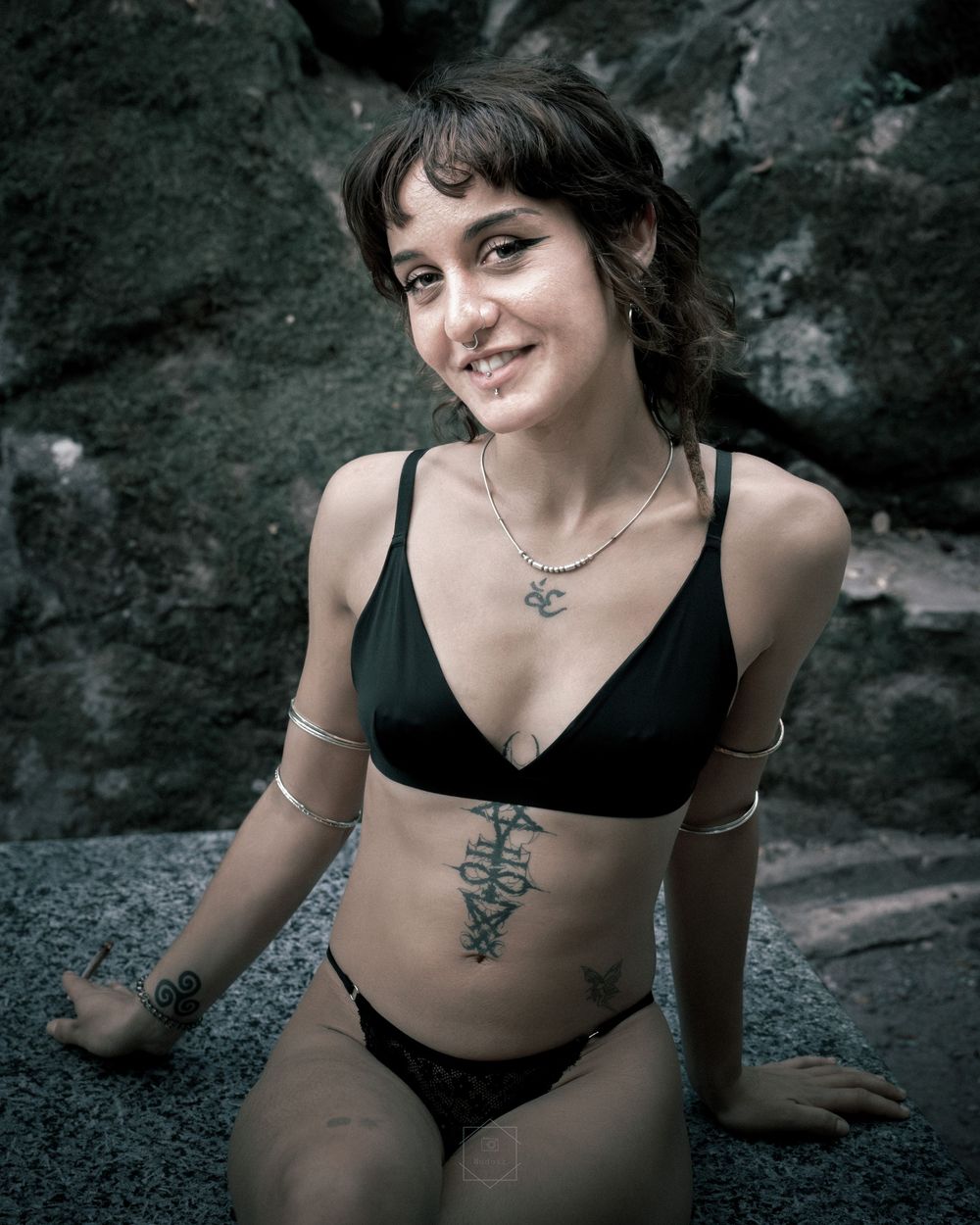 Young woman in black bikini with tattoos smiling outdoors near rocky background.