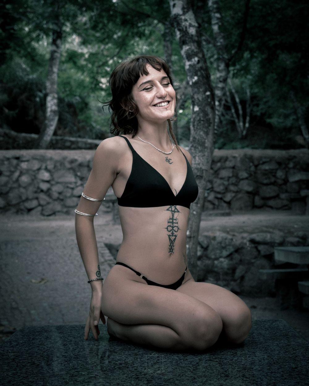 Smiling young woman in black bikini with side tattoo sitting outdoors near stone wall and trees.