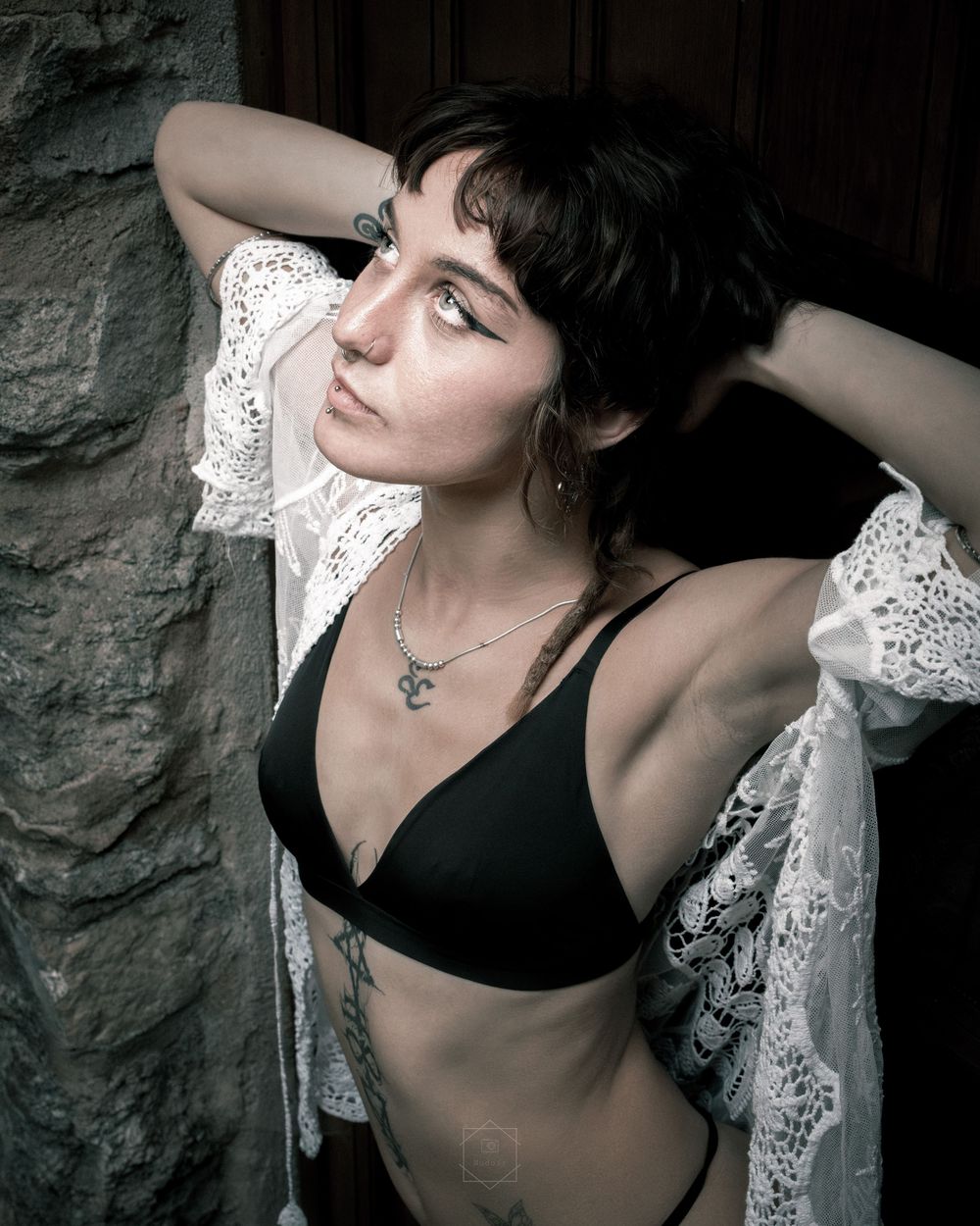 Young woman with short dark hair poses in black bikini top and white lace robe against stone wall with tattoos visible.