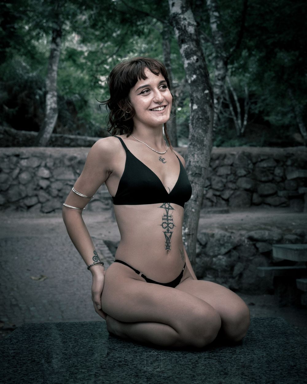 Young woman in black bikini with side tattoo sitting outdoors, smiling, with stone wall and trees in background.