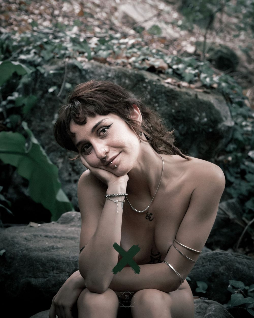 Young woman with curly hair poses outdoors near rocks and foliage, wearing jewelry and a green X body paint.
