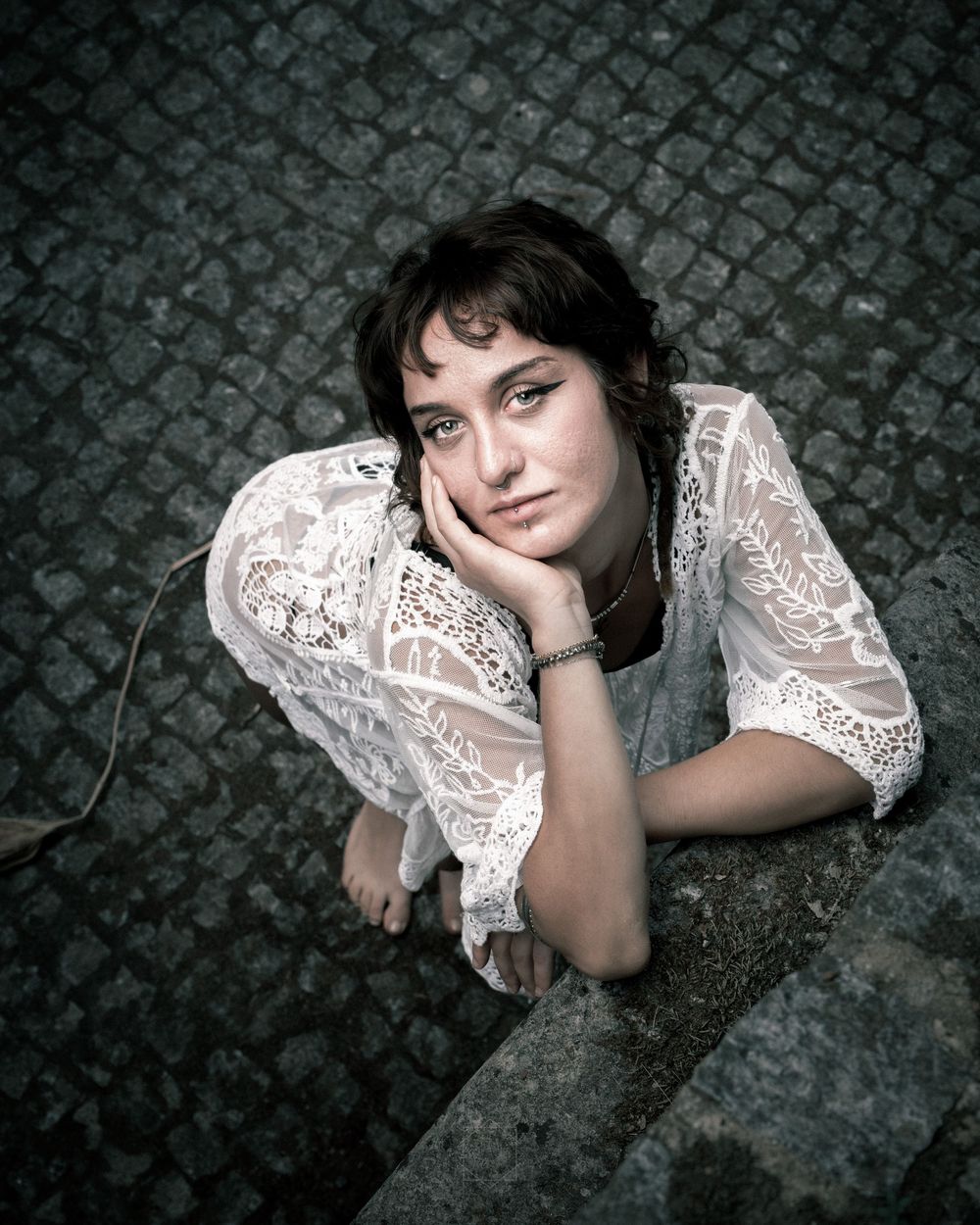 Young woman in white lace blouse resting chin on hand, sitting on stone steps with cobblestone background.