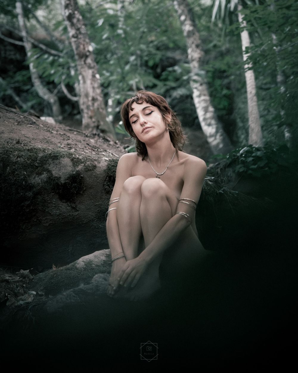 A woman sits peacefully on rocks in a lush forest, eyes closed, wearing gold jewelry.