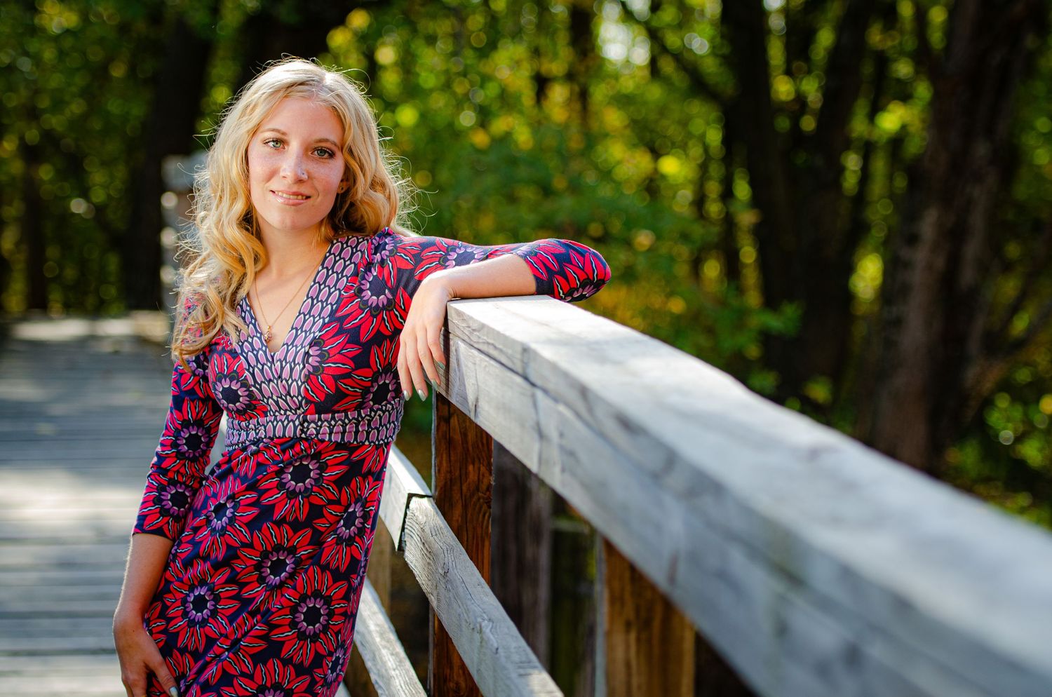 Blonde high school senior in sun dress leans against bridge railing smiling.