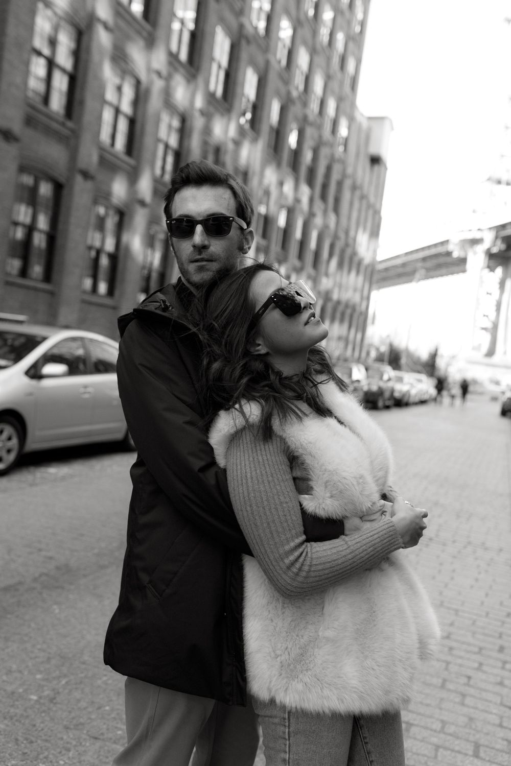 Black and white photo of a stylish couple embracing on a city street, both wearing sunglasses and fashionable coats.