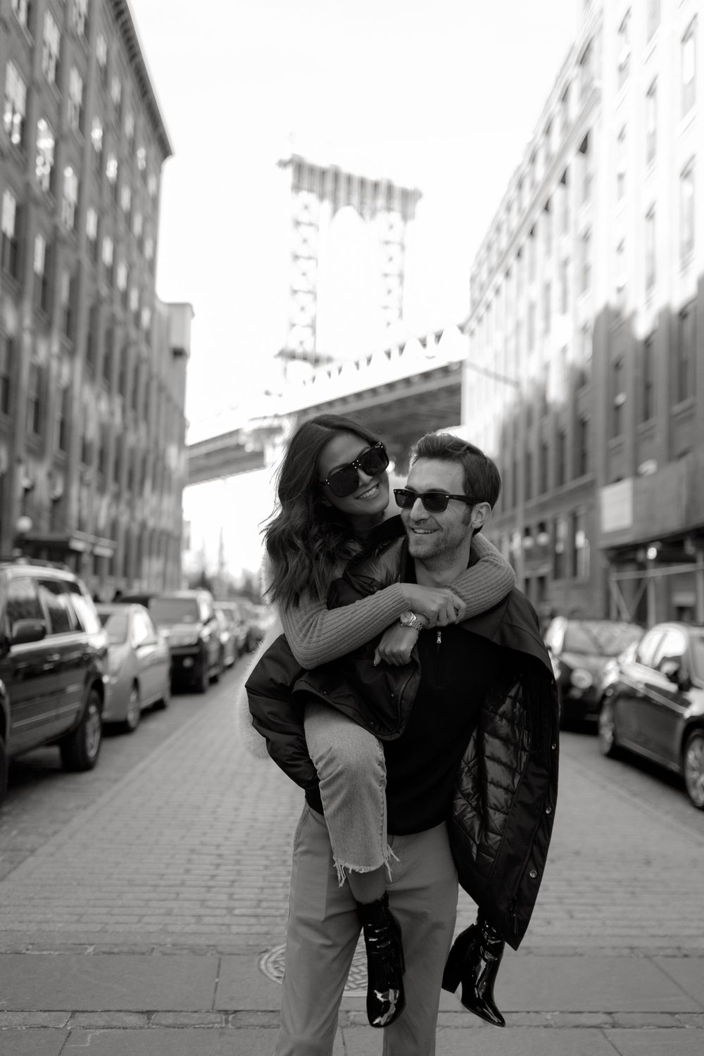 Black and white photo of a couple, man giving woman a piggyback ride on a city street with Manhattan Bridge in background.
