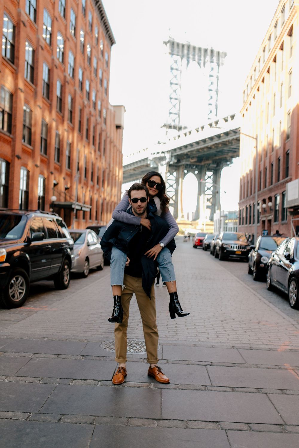Couple posing on a Brooklyn street, man giving woman a piggyback ride with Manhattan Bridge in background.