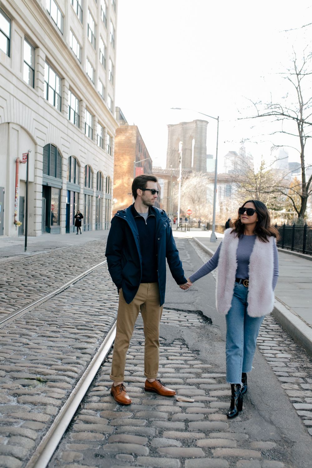 Stylish couple holding hands on cobblestone street near Brooklyn Bridge on a sunny day.