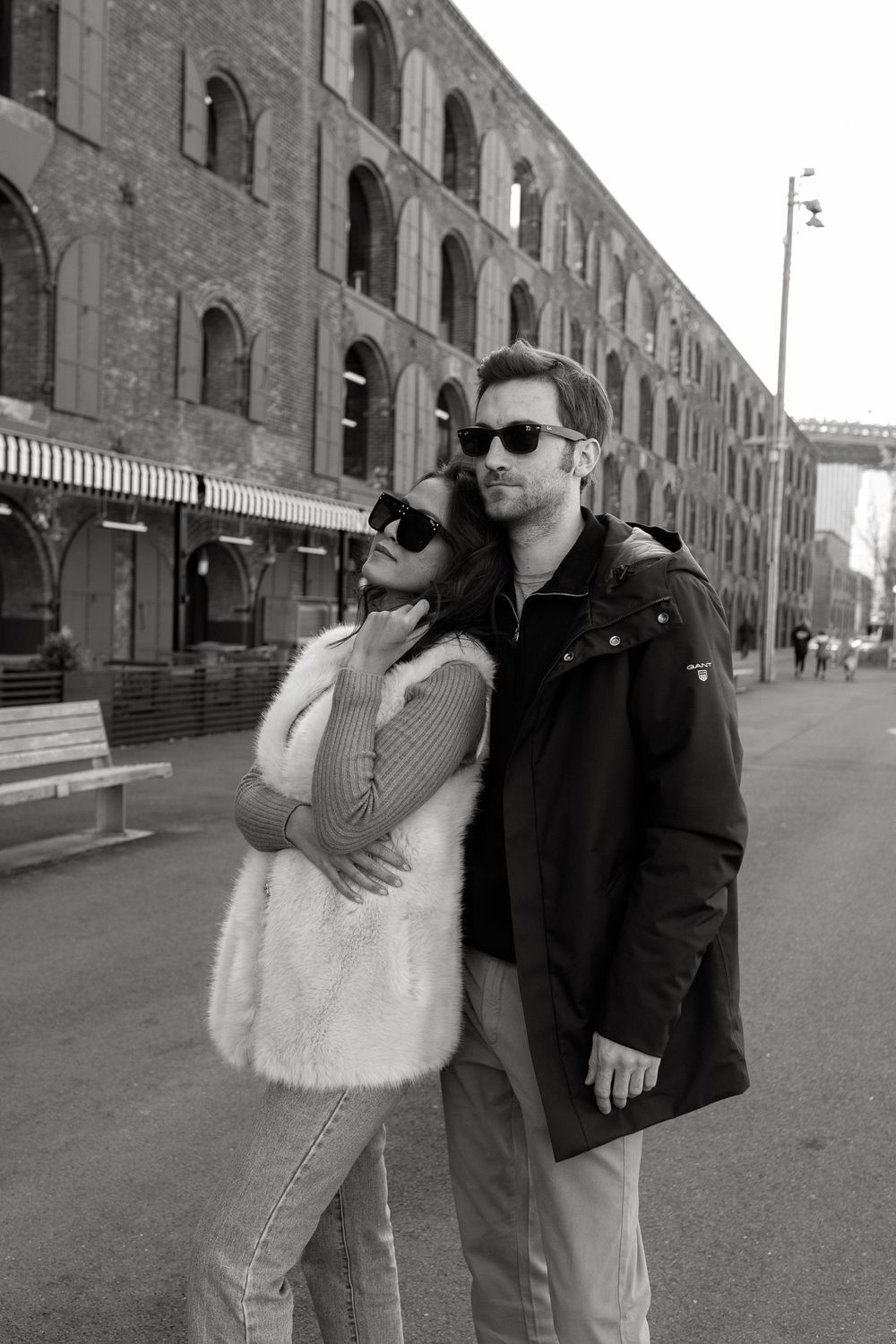 Stylish couple embracing on a city street in black and white, wearing sunglasses near a historic brick building.
