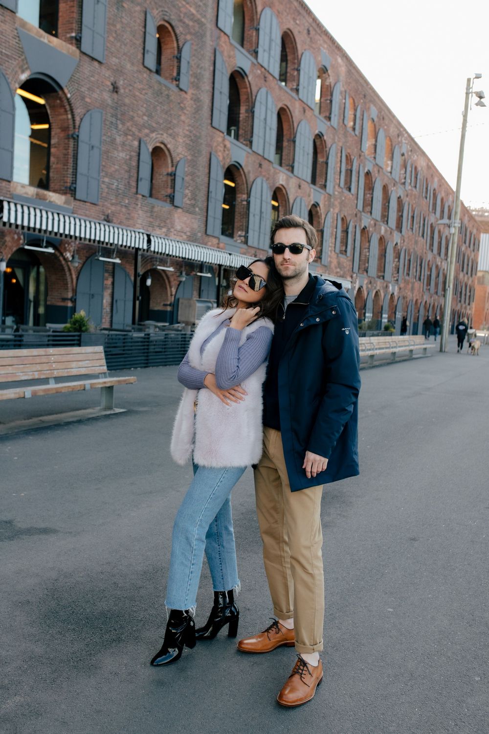Stylish couple posing on urban street near brick building, woman in fur vest and jeans, man in navy coat.