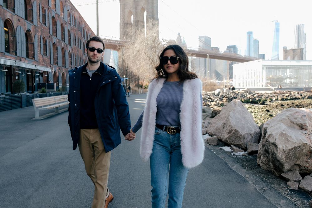 Couple holding hands walking near Brooklyn Bridge waterfront, woman in white fur vest and jeans.