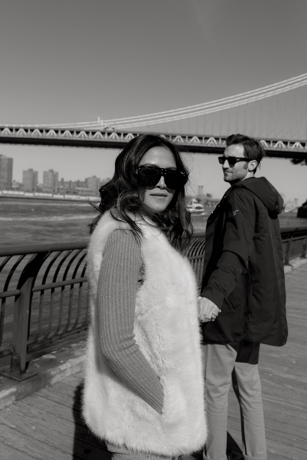 Black and white photo of woman in fur vest and sunglasses with man near Manhattan Bridge waterfront.