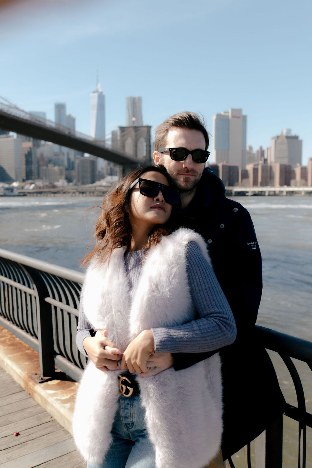 Couple in sunglasses posing on Brooklyn Bridge Park waterfront with NYC skyline and Brooklyn Bridge in background.