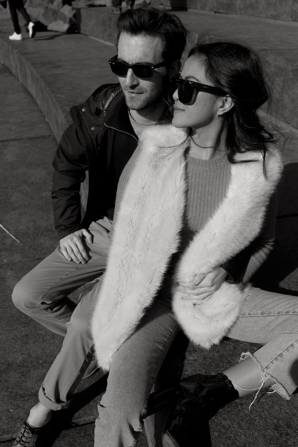 Black and white photo of a stylish couple wearing sunglasses and fur vest sitting outdoors in urban setting.