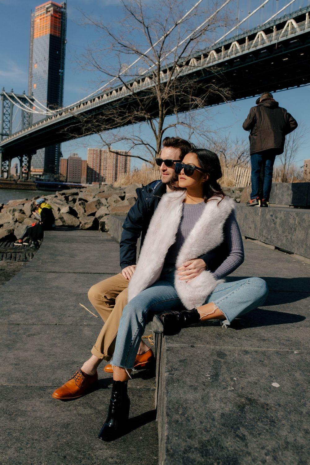 Couple sitting together near Manhattan Bridge in NYC, wearing sunglasses and stylish fall outfits on a sunny day.