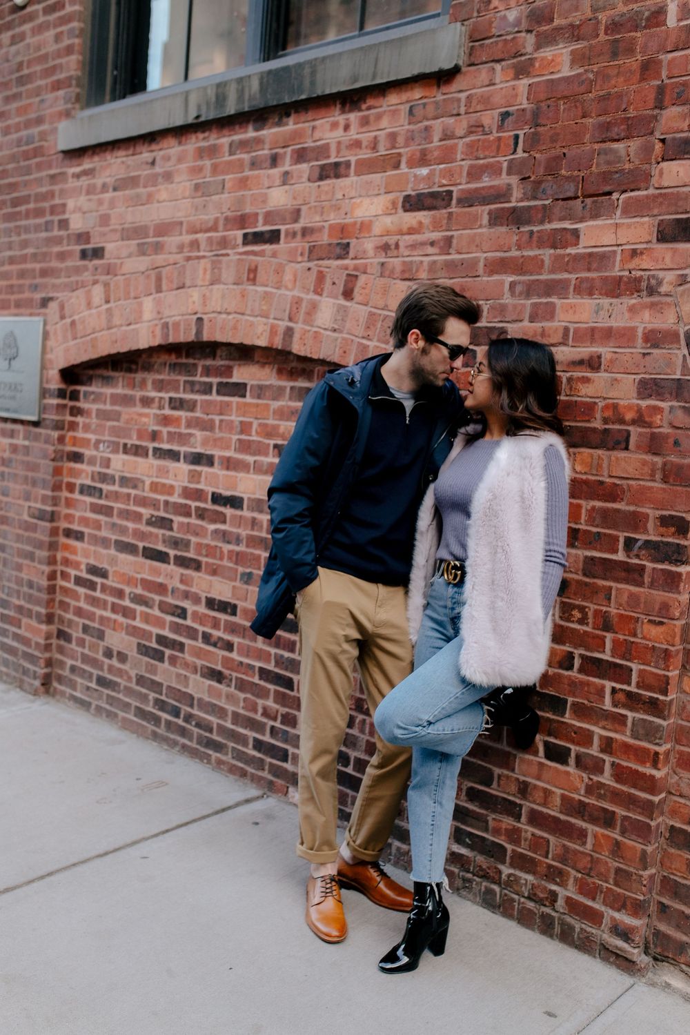 Couple sharing a romantic moment leaning against a red brick wall outdoors, dressed in stylish casual fall outfits.