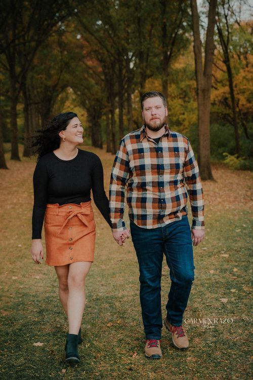 Couple holding hands walking through a park with autumn foliage, woman in orange skirt and man in plaid shirt.