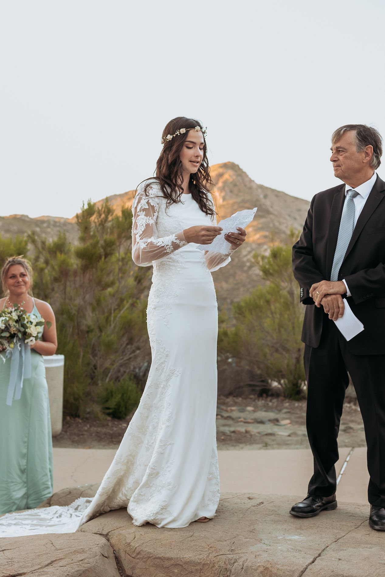 Bride in lace long-sleeve wedding dress reads vows outdoors at desert ceremony with bridesmaid and officiant nearby at Mission Trails.