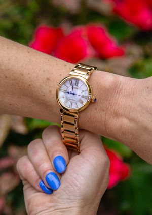 Gold watch with white face displayed on wrist with bright blue nails and red flowers in background.