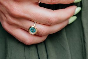 Close-up of hand wearing gold ring with teal gemstone against green fabric background.