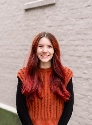 Woman in orange knit sweater with vibrant red hair smiles against neutral wall.