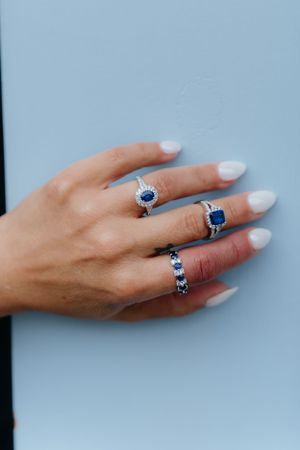 Hand wearing multiple sapphire and diamond rings against light blue background.