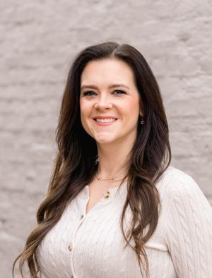 Woman in cream sweater smiles warmly against light colored brick wall background.