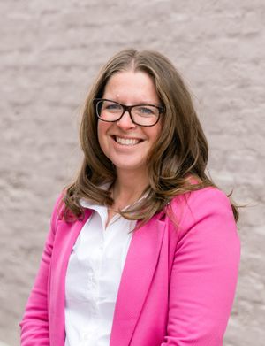 Professional headshot of a person wearing glasses, a pink blazer and white shirt against a neutral background.