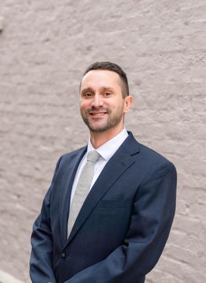 Professional headshot portrait of a person in a navy suit and gray tie against a light wall.