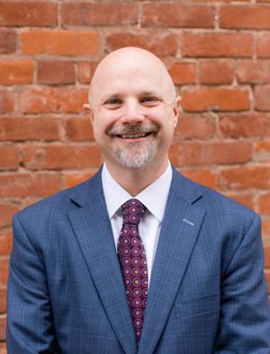 Professional portrait showing a person in a blue suit and patterned tie against a brick wall.
