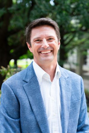 Professional headshot of a person in a blue blazer and white shirt against greenery.