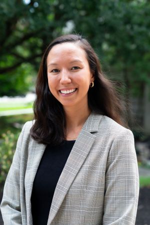 Professional headshot of a person in a plaid blazer against a natural outdoor background.