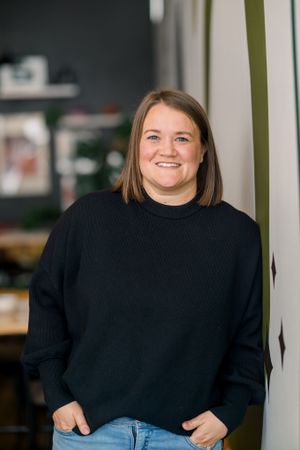 Person in black sweater and jeans smiling warmly in front of blurred office background.